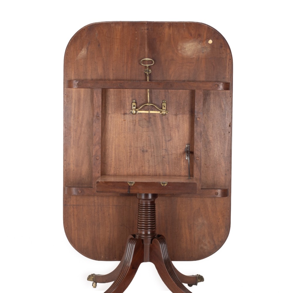 A George III mahogany dining table featuring a rectangular tilt-top surface with rounded corners set on a reeded central pedestal and four splayed legs with brass paw castors. View XI