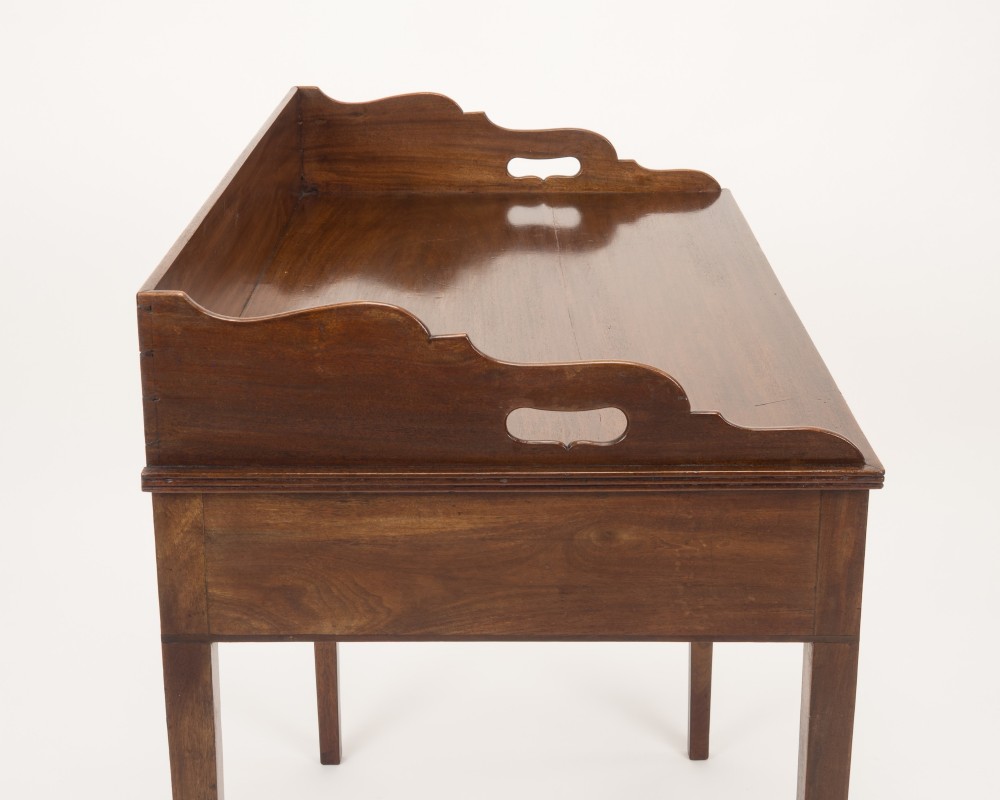 An early 19th-century mahogany tray-top writing table featuring a gallery edge with side cutout handles, two drawers with knob handles, and square tapered legs. View X