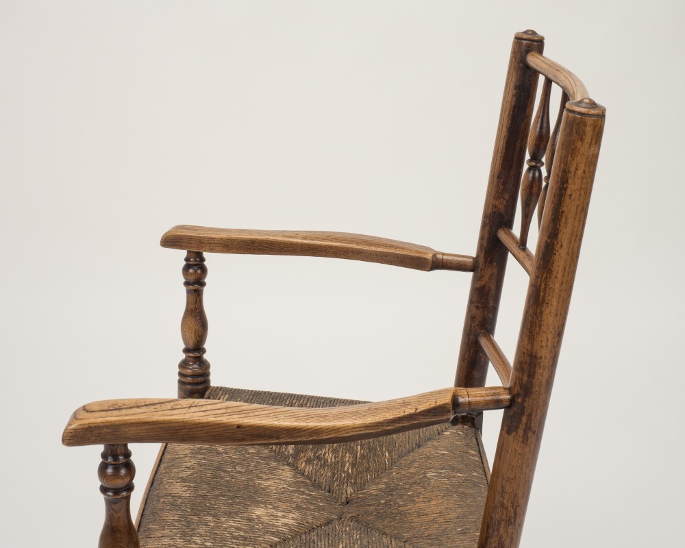 An early nineteenth-century English country armchair with a spindle back rest, curved open armrests, turned legs, and a woven rush seat. View V