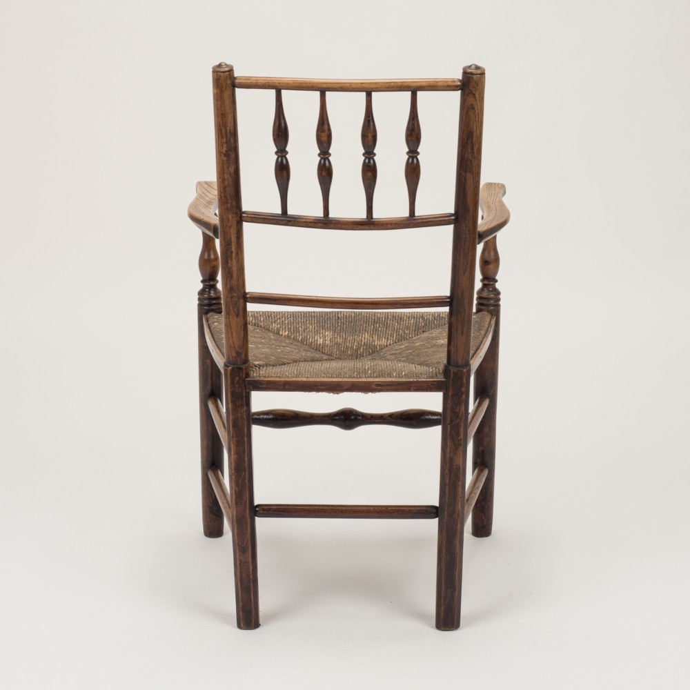 An early nineteenth-century English country armchair with a spindle back rest, curved open armrests, turned legs, and a woven rush seat. View X