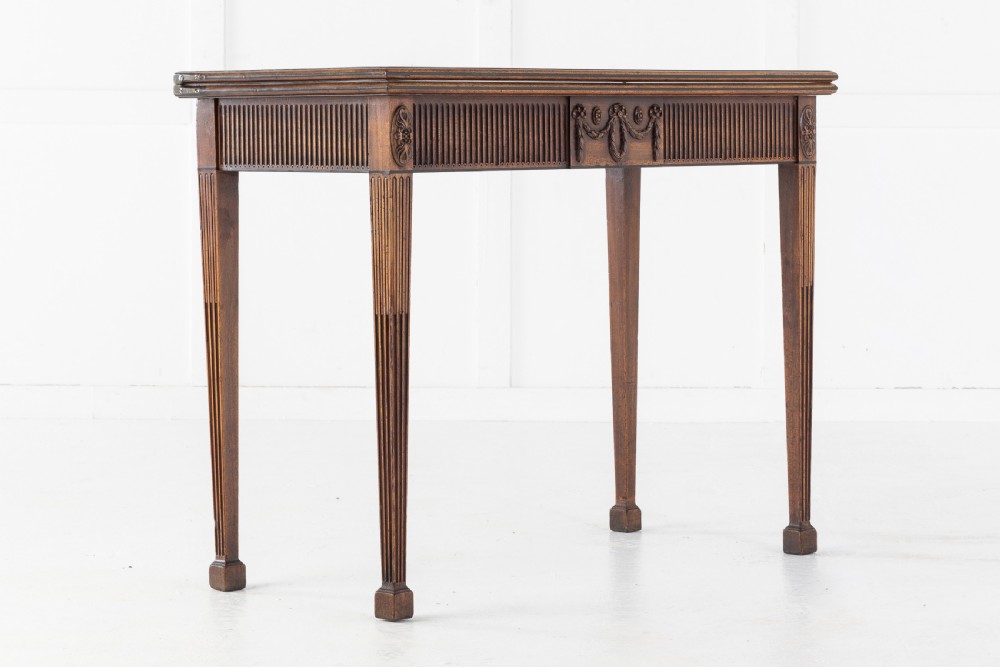 An 18th-century George III mahogany tea table featuring fluted tapering square legs, a reeded frieze with central carved floral swags, and a fold-over top.
