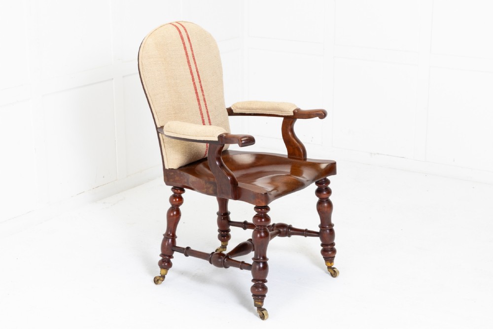 A 19th-century mahogany armchair featuring turned legs on brass castors, a contoured wooden saddle seat, padded armrests, and a beige linen upholstered backrest with central red stripes.