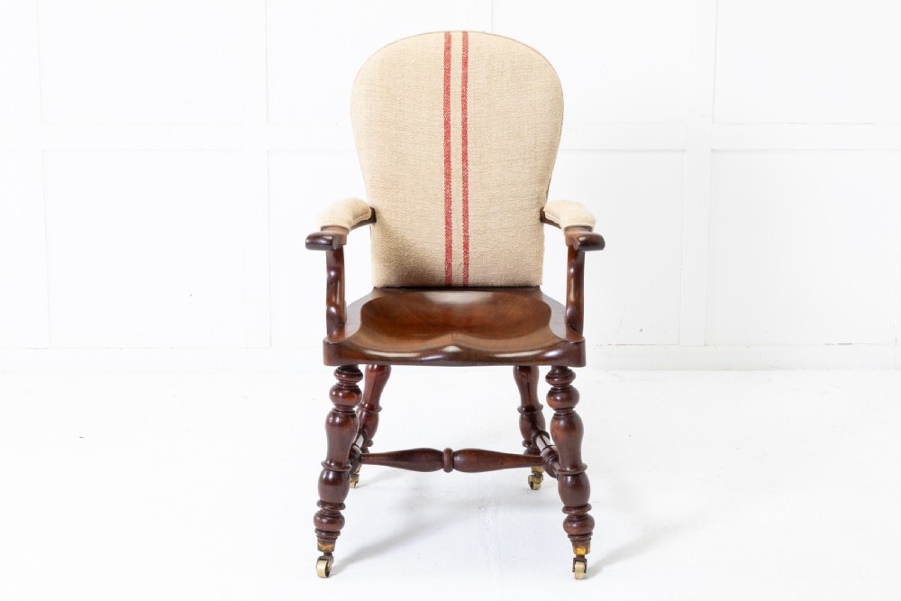 A 19th-century mahogany armchair featuring turned legs on brass castors, a contoured wooden saddle seat, padded armrests, and a beige linen upholstered backrest with central red stripes. View II