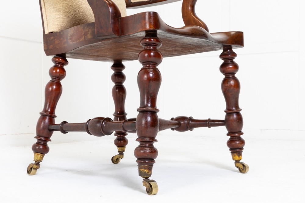 A 19th-century mahogany armchair featuring turned legs on brass castors, a contoured wooden saddle seat, padded armrests, and a beige linen upholstered backrest with central red stripes. View IV