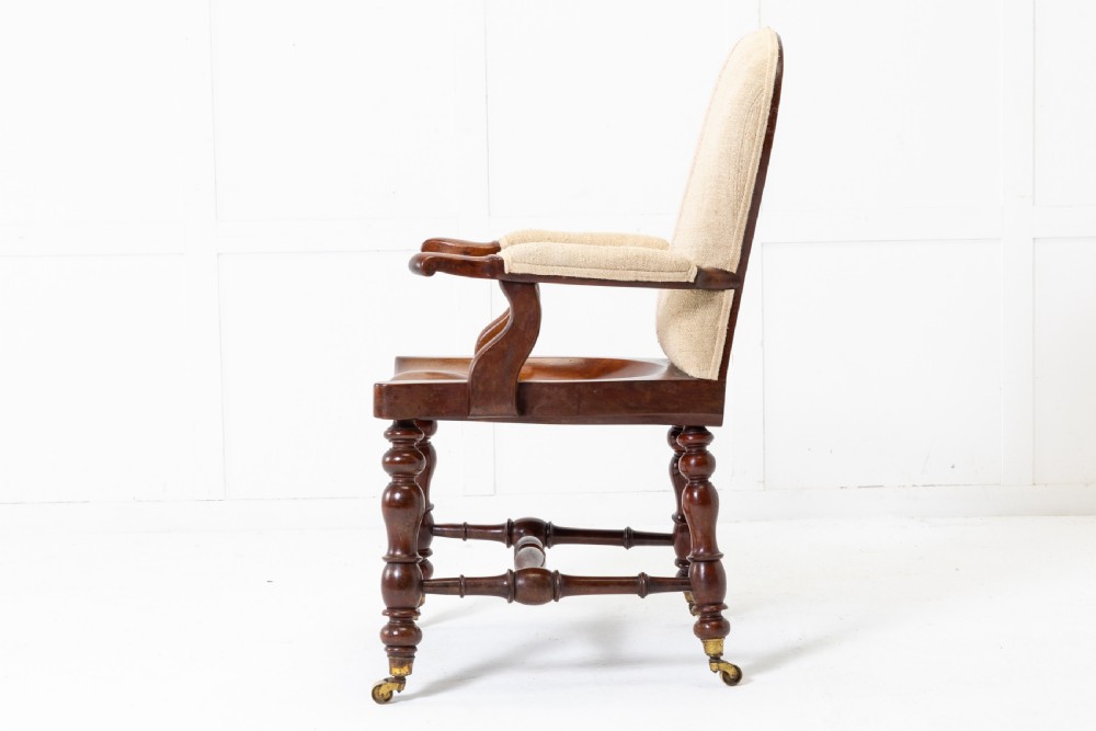 A 19th-century mahogany armchair featuring turned legs on brass castors, a contoured wooden saddle seat, padded armrests, and a beige linen upholstered backrest with central red stripes. View V