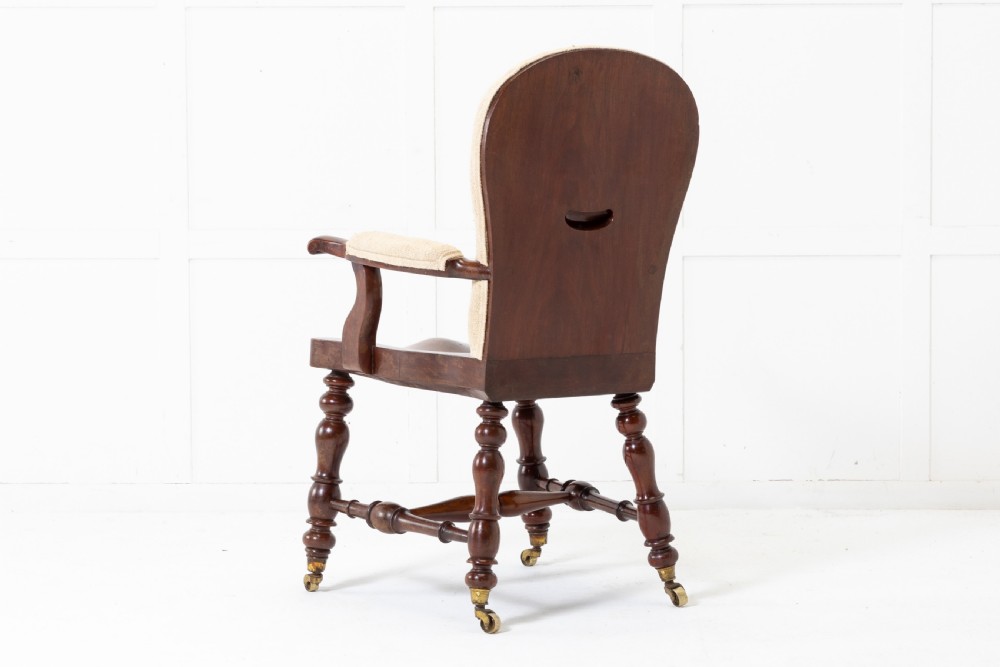 A 19th-century mahogany armchair featuring turned legs on brass castors, a contoured wooden saddle seat, padded armrests, and a beige linen upholstered backrest with central red stripes. View VII