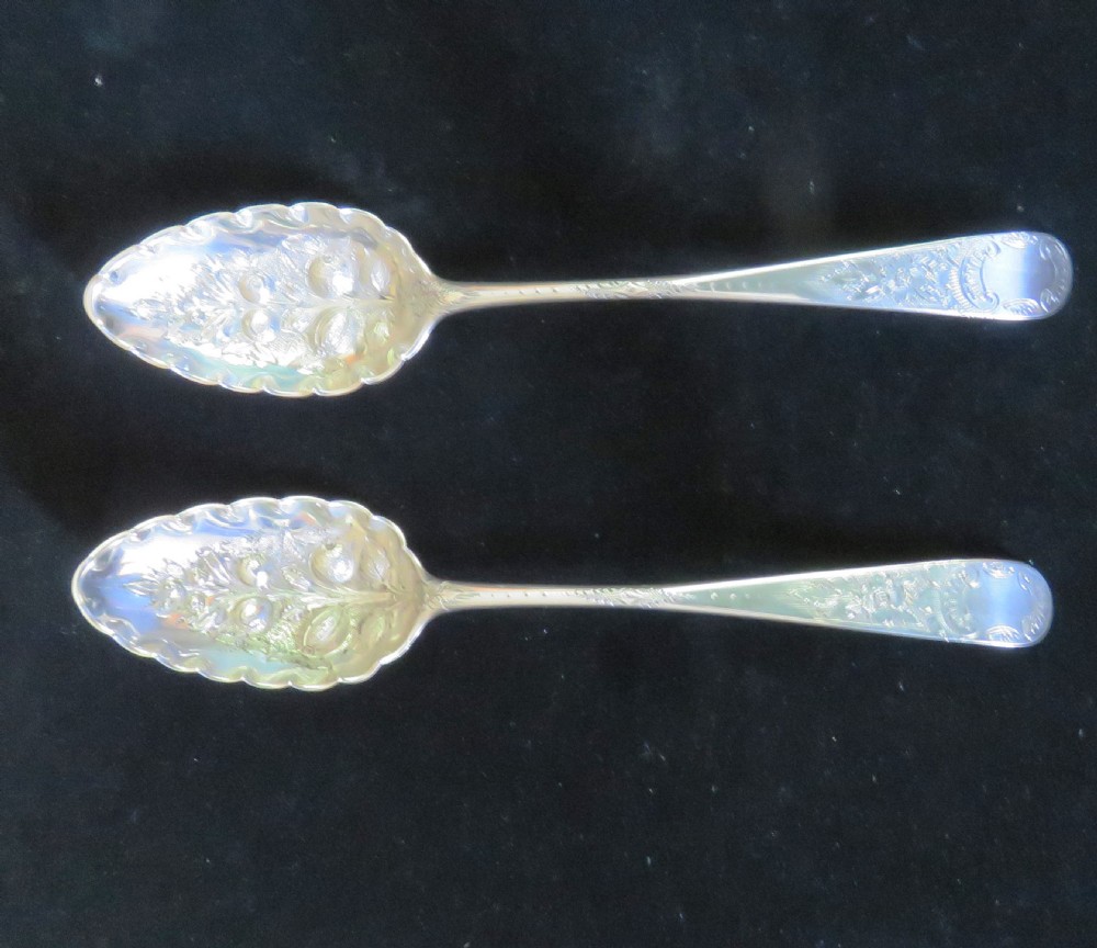 A pair of Georgian sterling silver berry spoons featuring repousse fruit motifs on shaped bowls and detailed engraving along the handles.