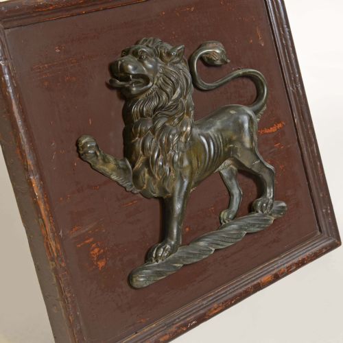 fig 2. A cast bronze lion passant heraldic relief mounted on a rectangular dark red-brown painted wooden plaque.