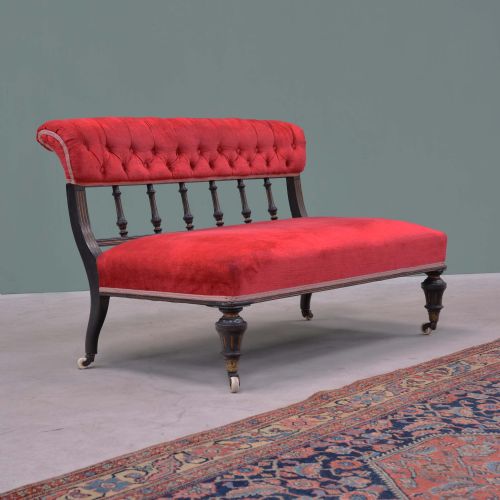 fig 1. An Aesthetic Movement ebonised wooden settee featuring red velvet button-back upholstery, turned spindle supports, and turned legs on ceramic castors.