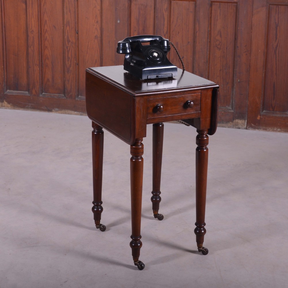 A dark mahogany drop-leaf occasional table featuring a single drawer with two round wooden knobs, four turned legs, and brass castors.