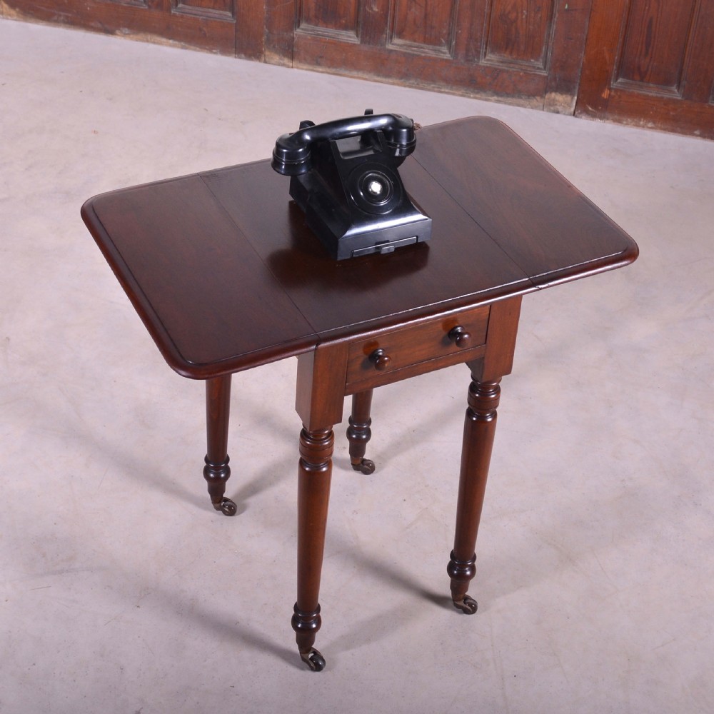 A dark mahogany drop-leaf occasional table featuring a single drawer with two round wooden knobs, four turned legs, and brass castors. View II