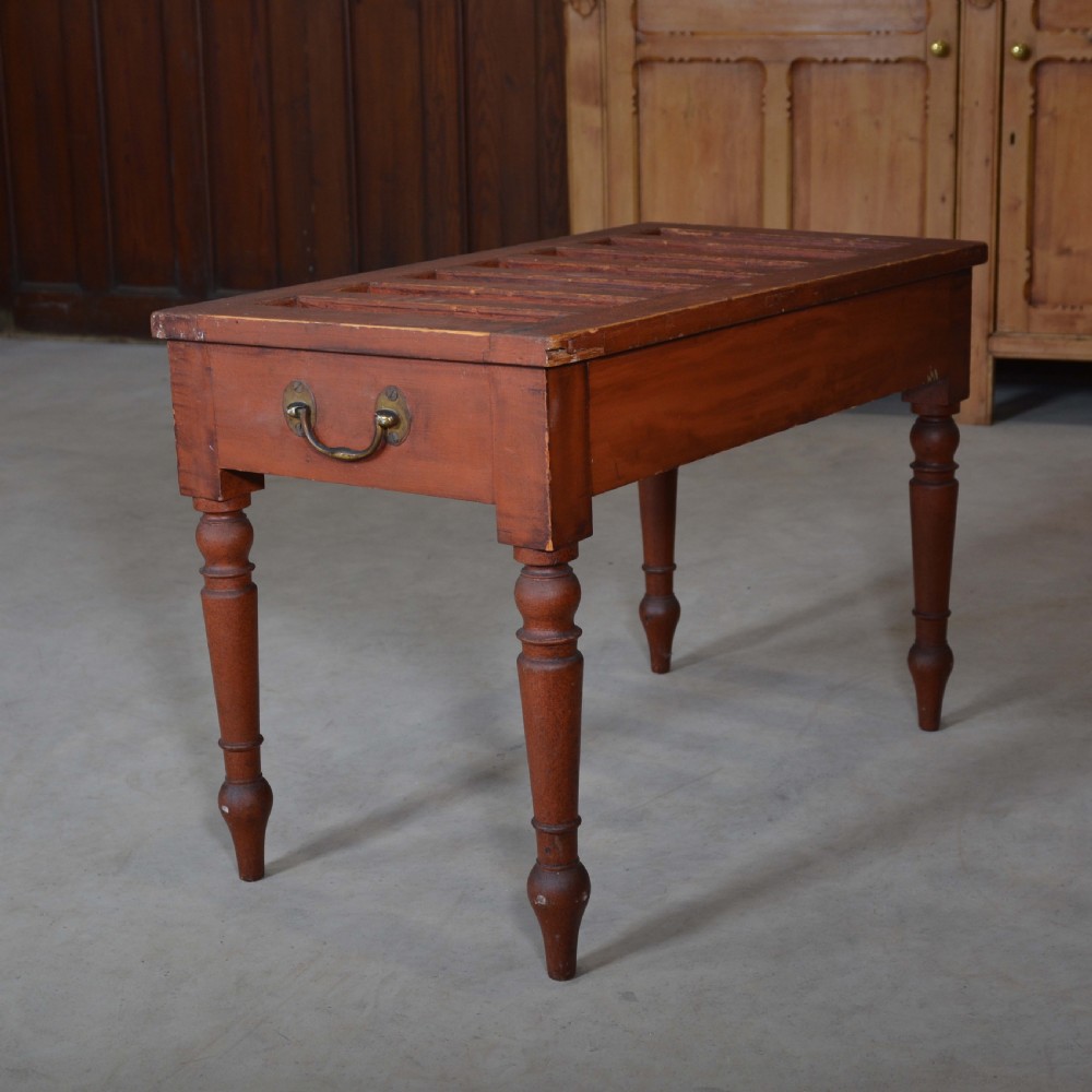 A low rectangular pine luggage stand painted in terracotta red with a slatted top surface, side handles, and four turned legs. View III