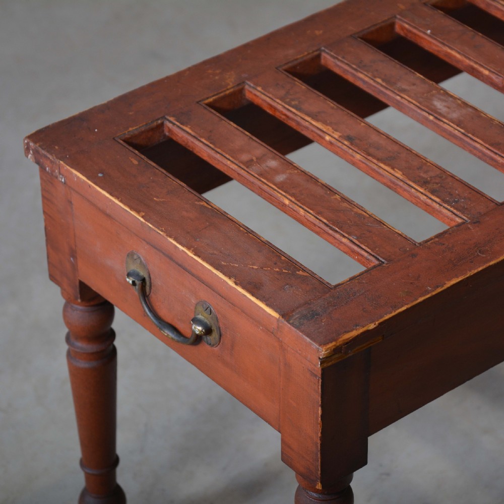 A low rectangular pine luggage stand painted in terracotta red with a slatted top surface, side handles, and four turned legs. View IV