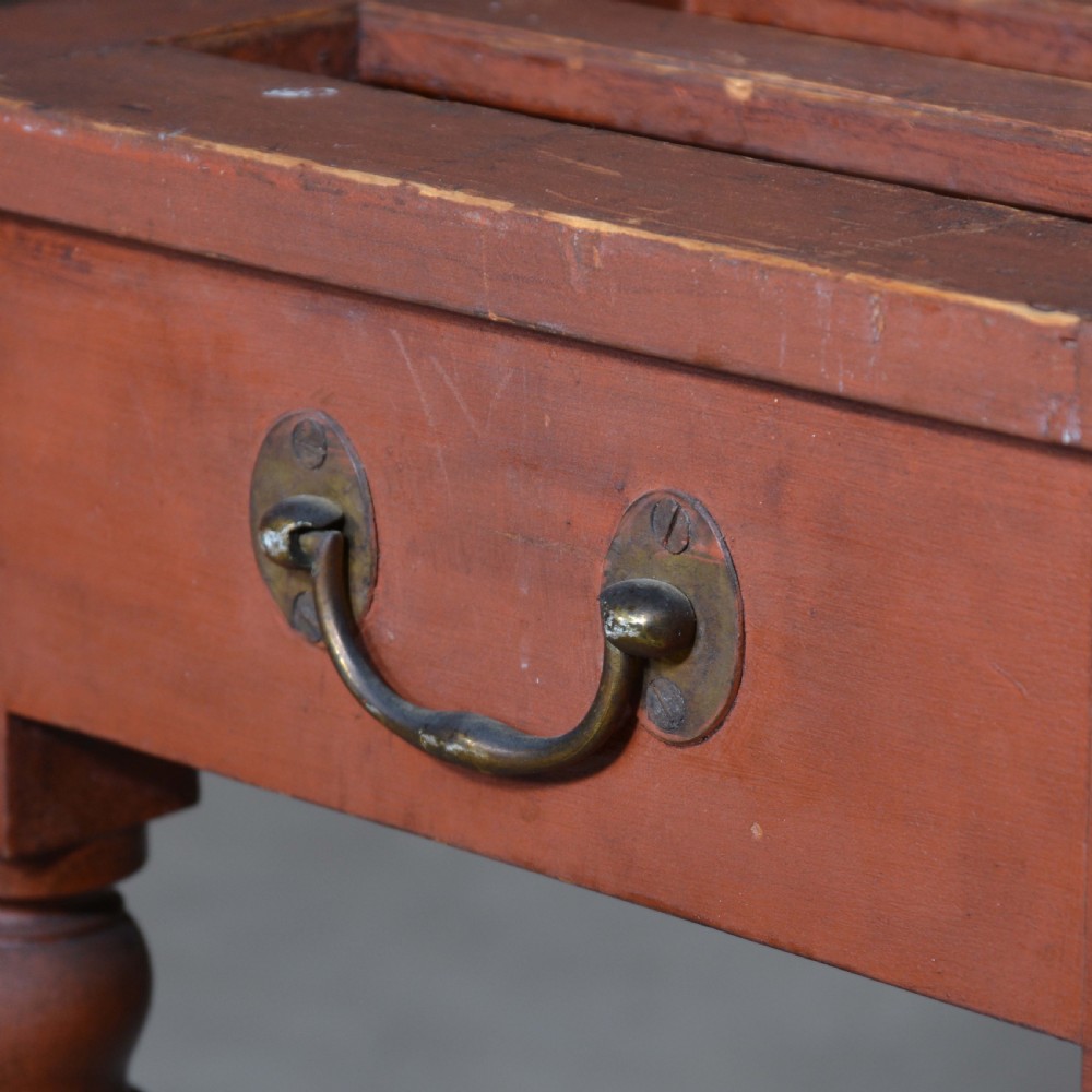 A low rectangular pine luggage stand painted in terracotta red with a slatted top surface, side handles, and four turned legs. View VI