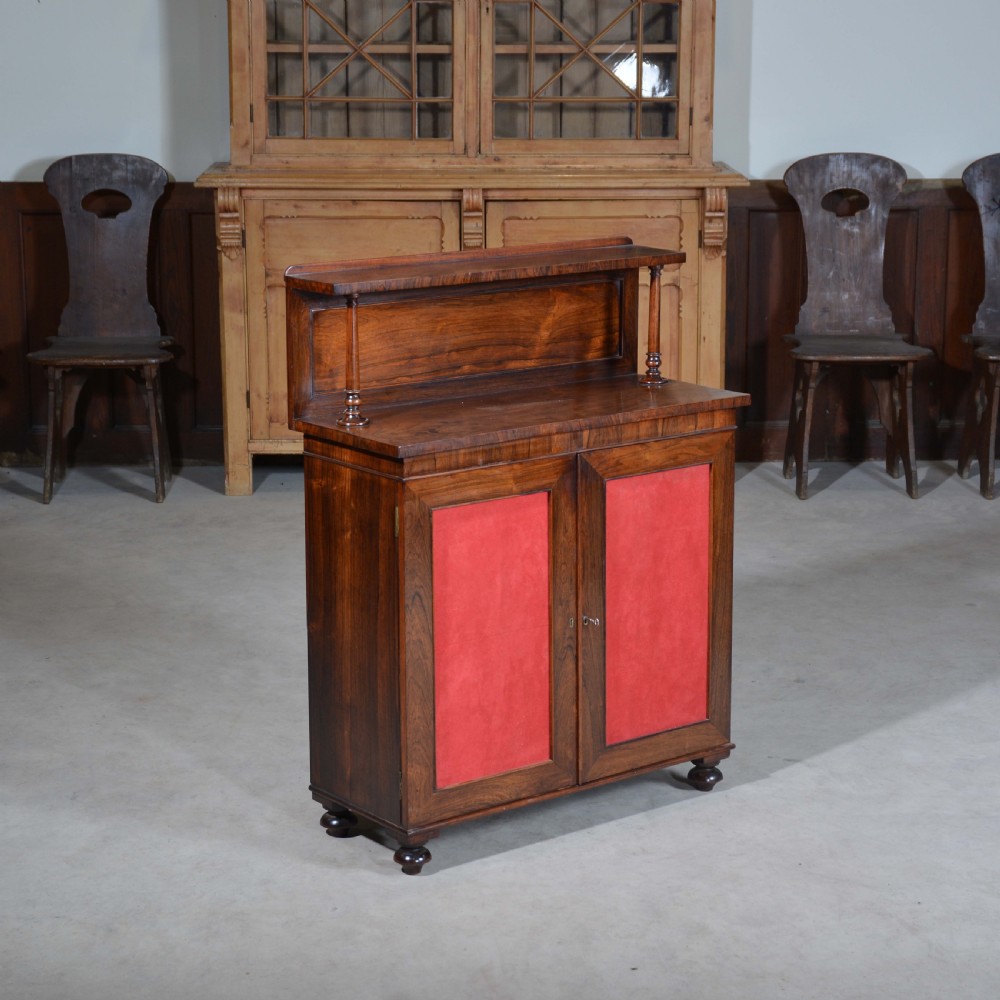 A Regency dwarf rosewood chiffonier featuring two red fabric-lined panel doors, a raised upper shelf supported by turned columns, and bun feet.