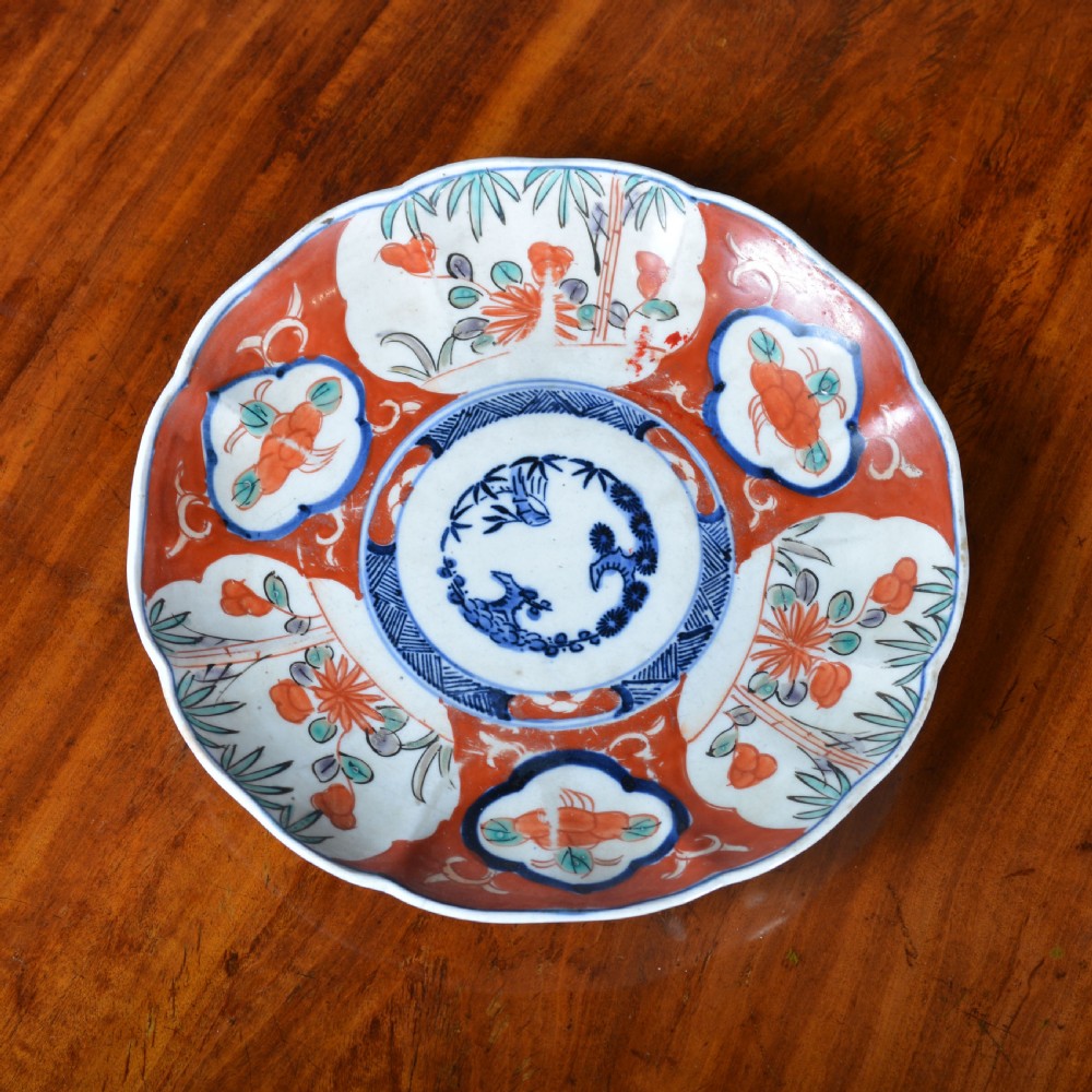 A 19th-century Japanese Imari porcelain circular dish featuring a scalloped rim, central blue medallion, and panelled floral decoration in iron red, cobalt blue, and green enamels.