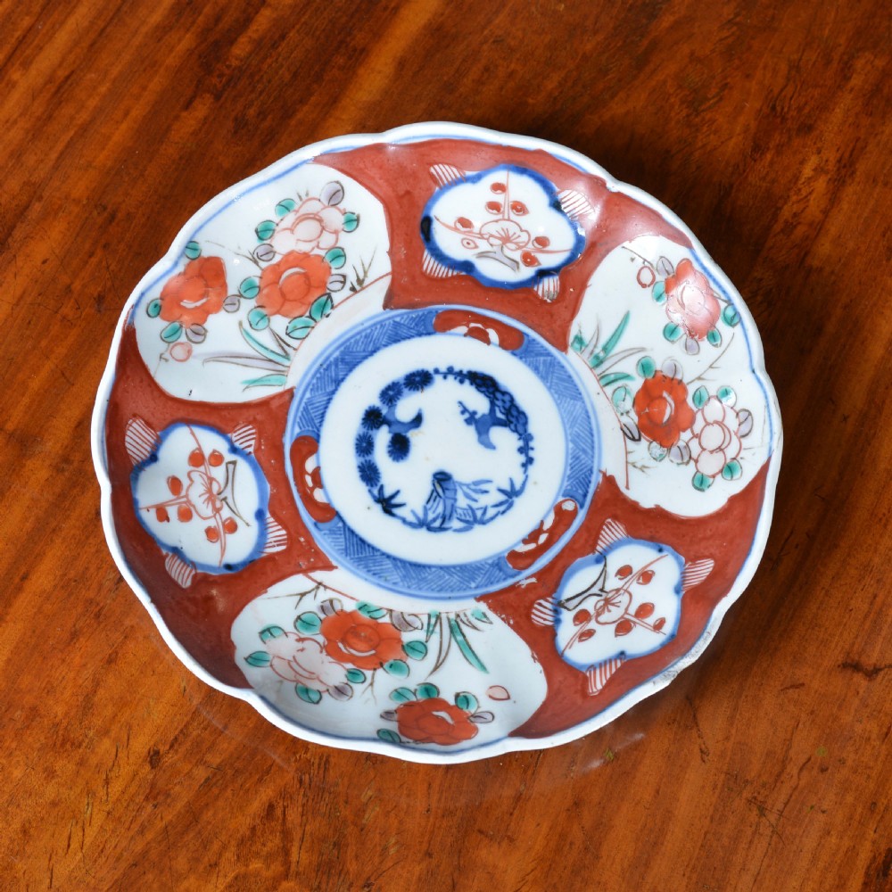 A circular 19th-century Japanese Imari porcelain plate featuring a scalloped rim, hand-painted rust-red and cobalt-blue floral motifs, and a central blue medallion.