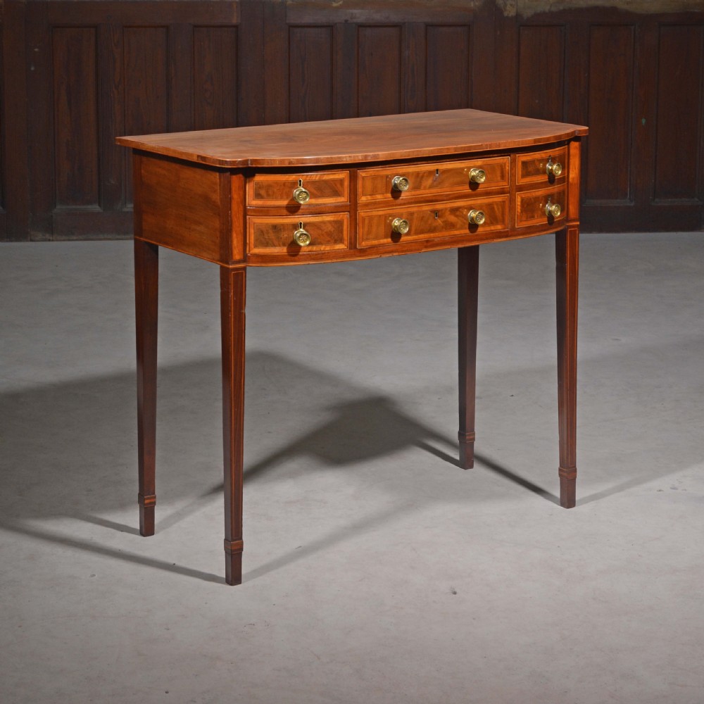 An antique Sheraton style mahogany bowfront side table with stringing inlay, brass handles, and square tapered legs.