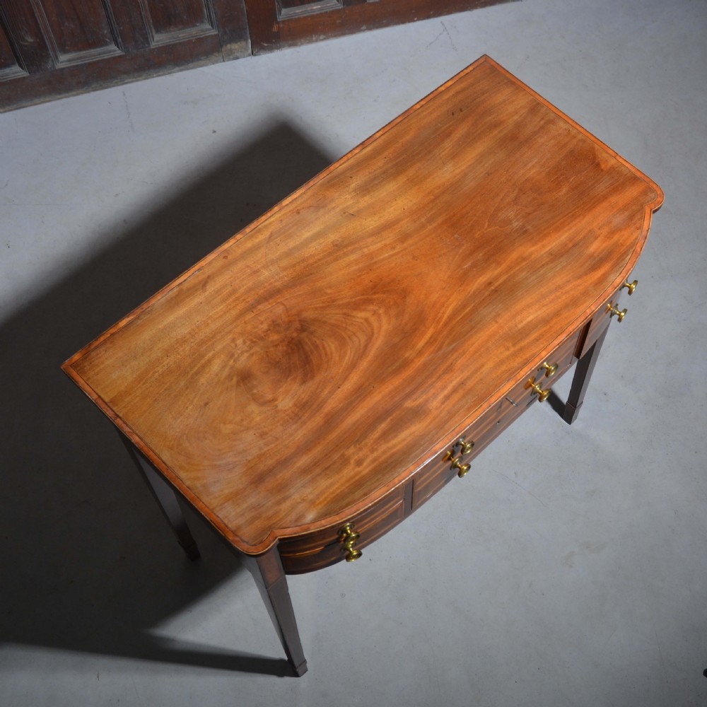 An antique Sheraton style mahogany bowfront side table with stringing inlay, brass handles, and square tapered legs. View X