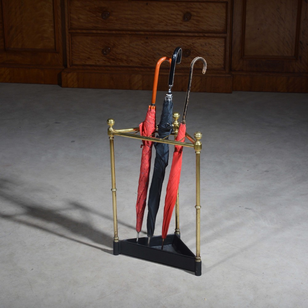 A triangular brass corner stick stand featuring tubular uprights topped with finials and a black cast iron drip tray base.