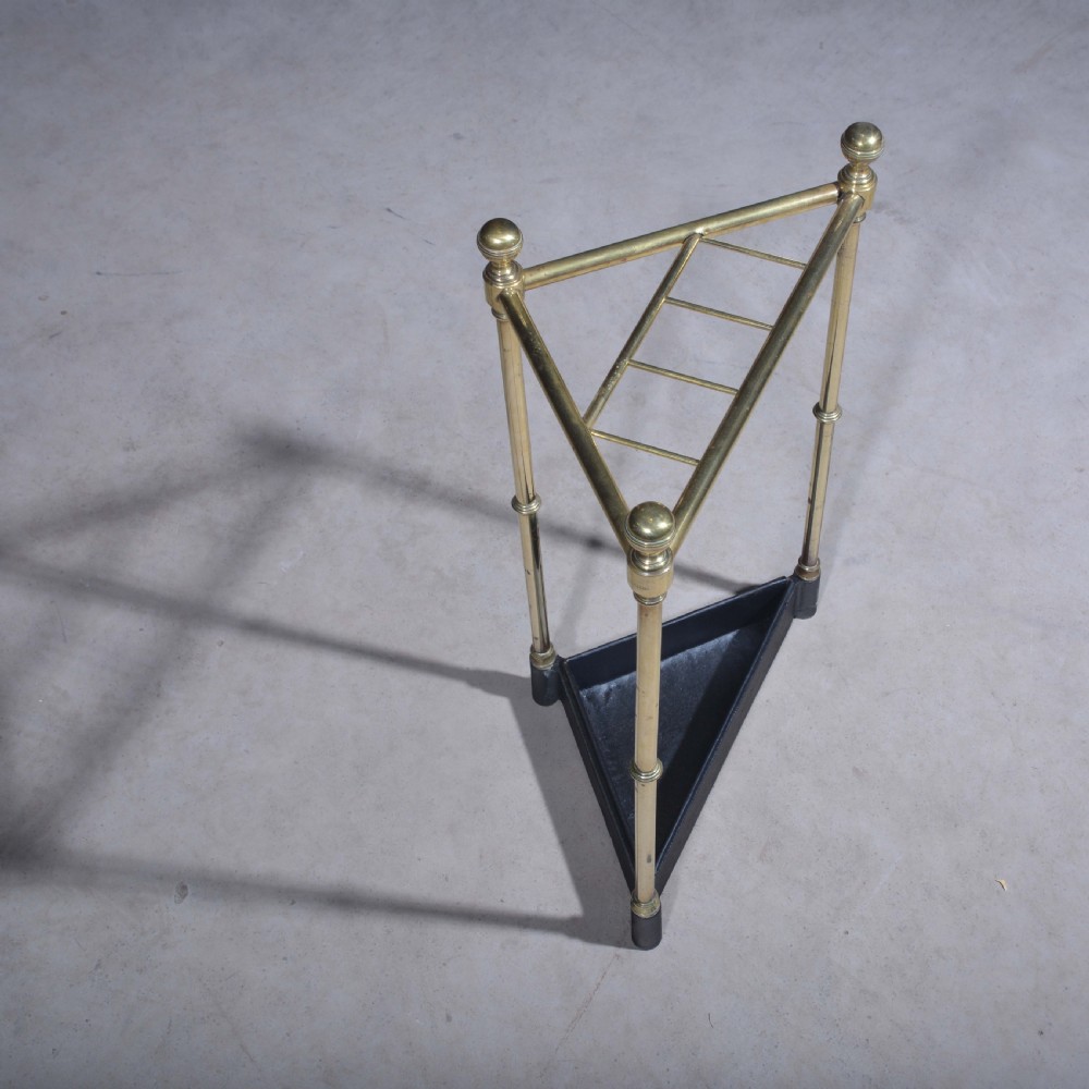 A triangular brass corner stick stand featuring tubular uprights topped with finials and a black cast iron drip tray base. View V