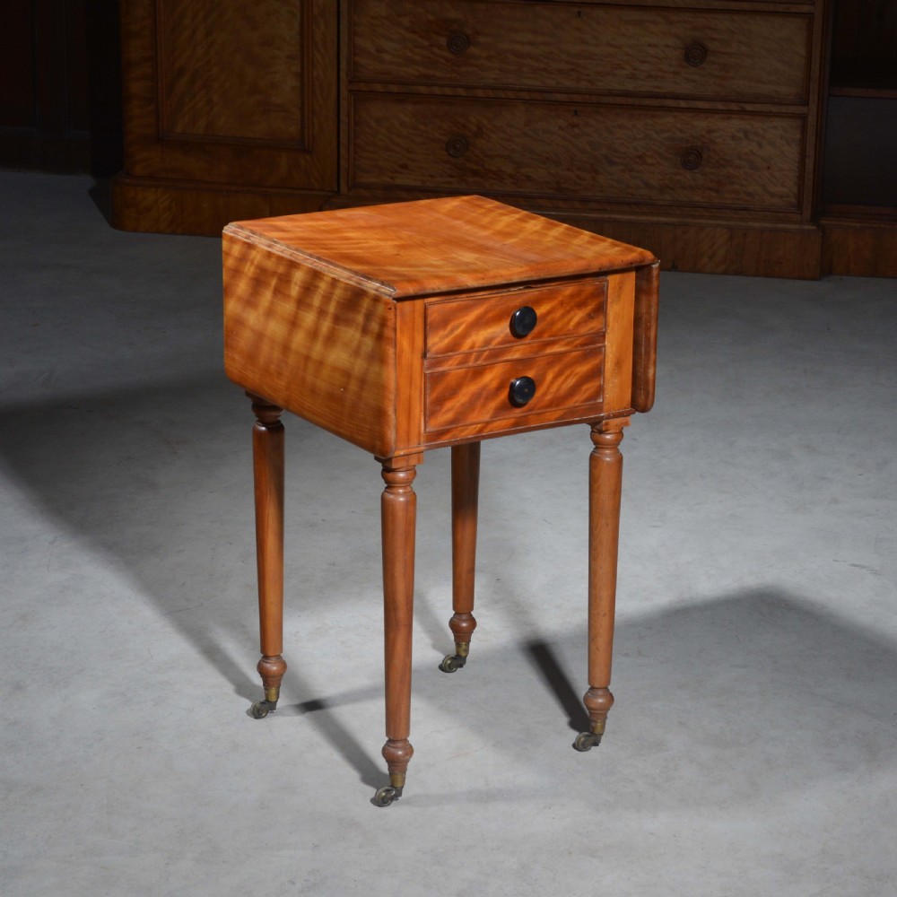 A 19th-century satin birch bedside table featuring a drop-leaf top, two small drawers with dark circular knobs, and turned legs terminating in brass castors.