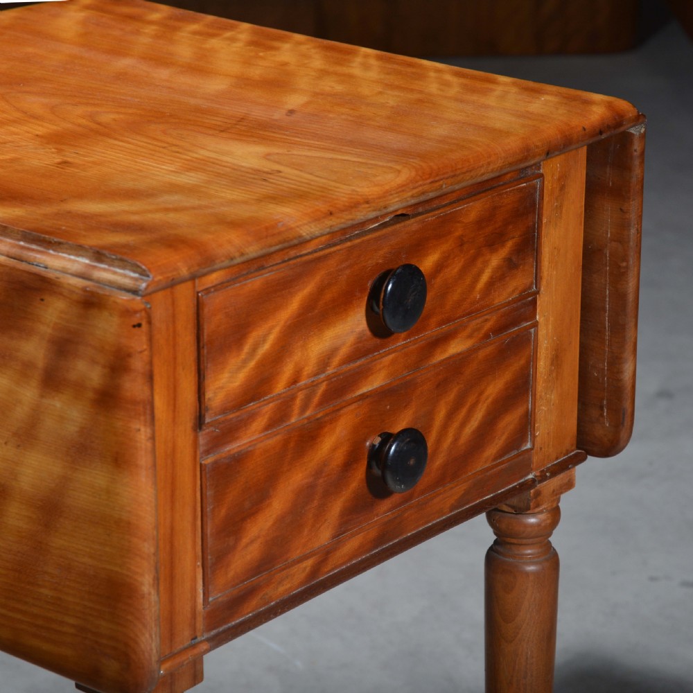 A 19th-century satin birch bedside table featuring a drop-leaf top, two small drawers with dark circular knobs, and turned legs terminating in brass castors. View II