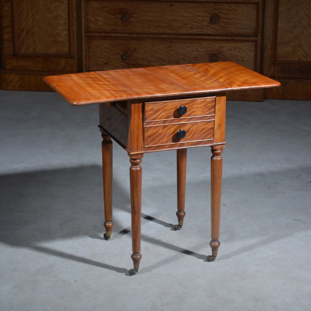 A 19th-century satin birch bedside table featuring a drop-leaf top, two small drawers with dark circular knobs, and turned legs terminating in brass castors. View III