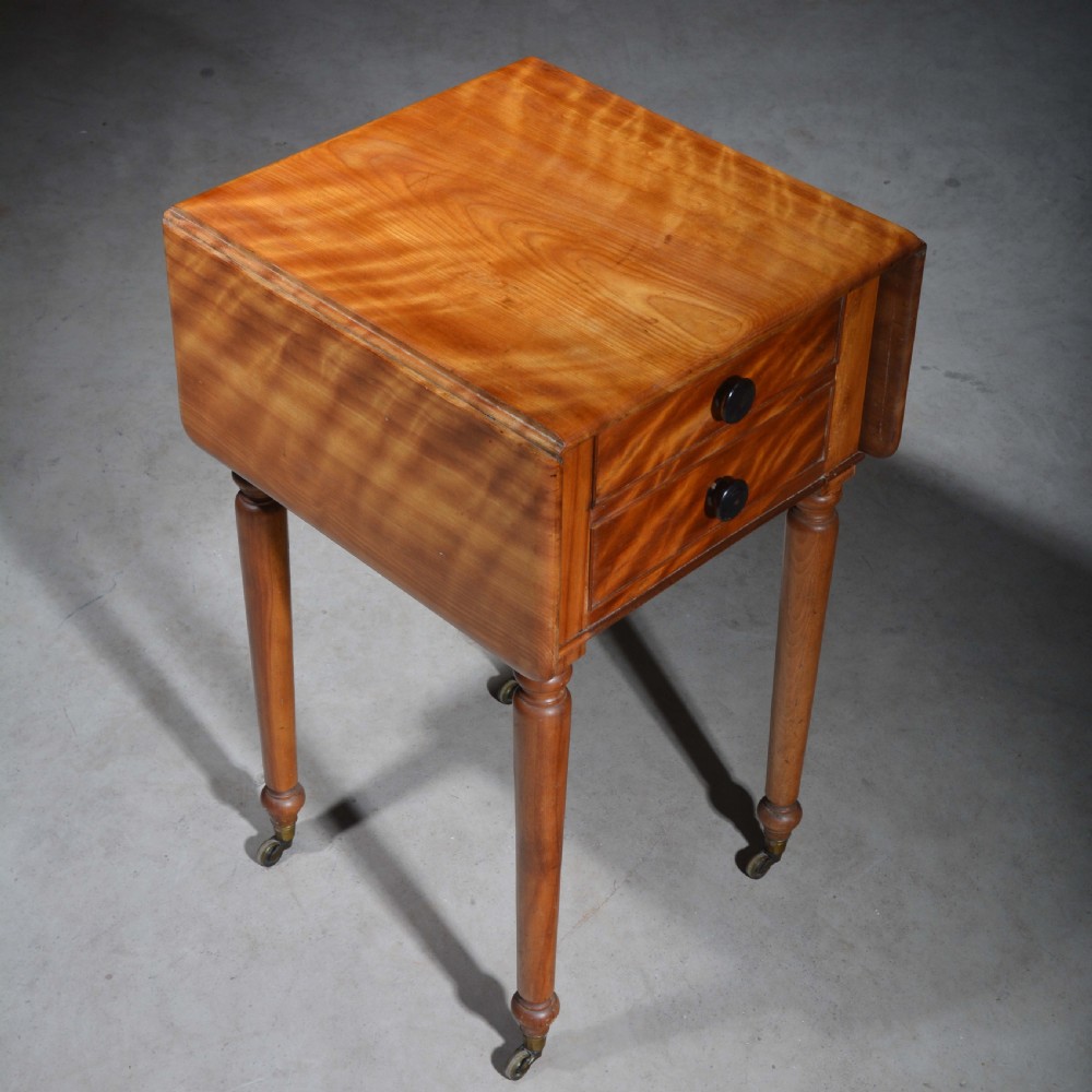 A 19th-century satin birch bedside table featuring a drop-leaf top, two small drawers with dark circular knobs, and turned legs terminating in brass castors. View IV
