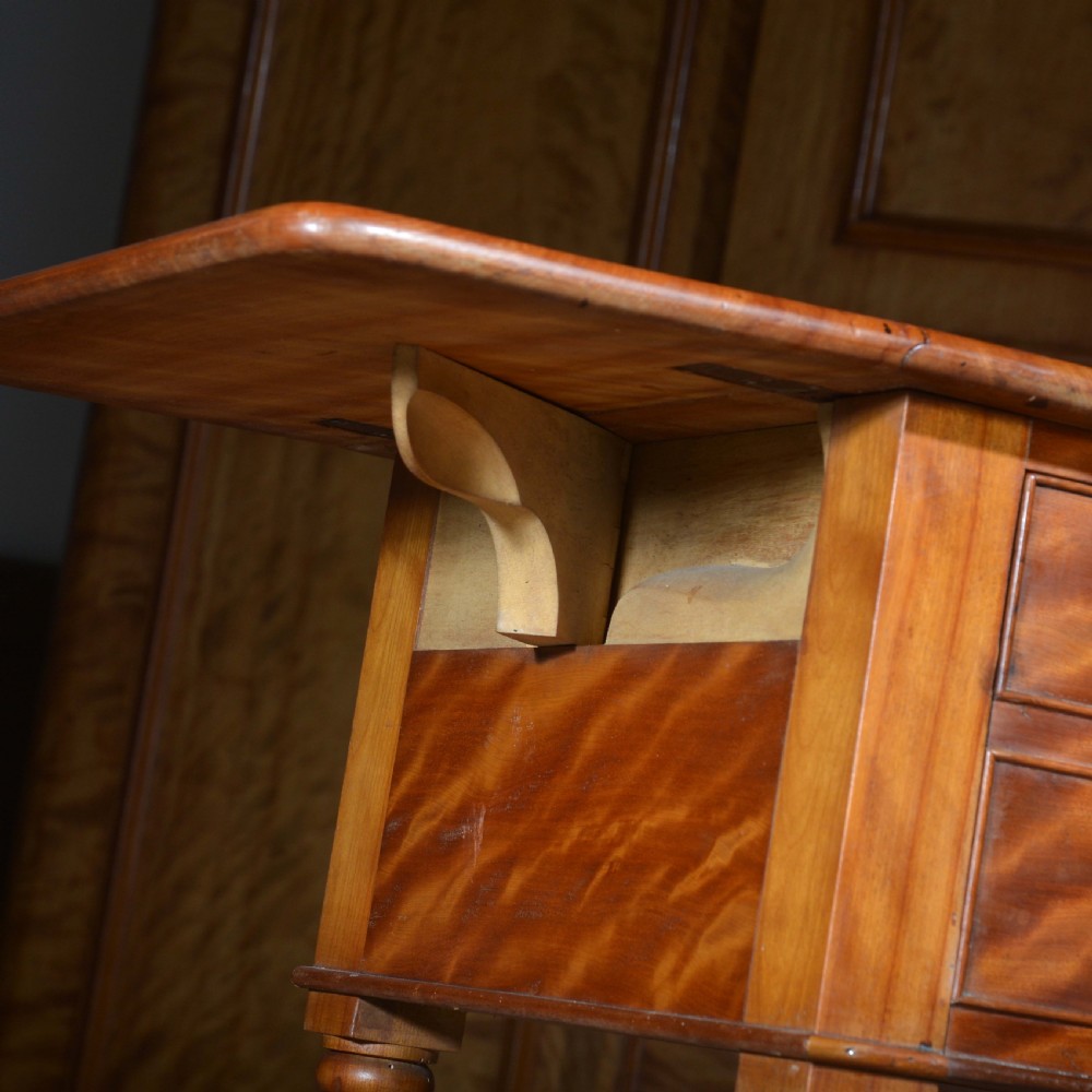 A 19th-century satin birch bedside table featuring a drop-leaf top, two small drawers with dark circular knobs, and turned legs terminating in brass castors. View V