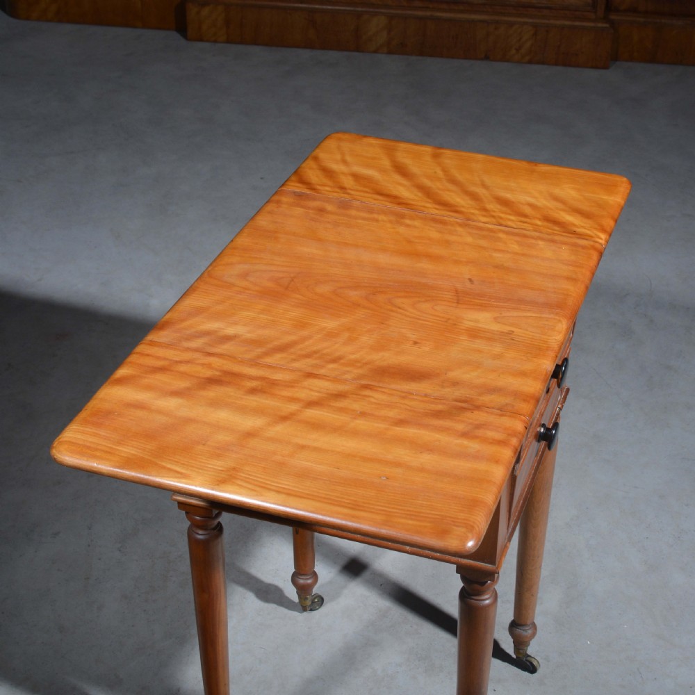 A 19th-century satin birch bedside table featuring a drop-leaf top, two small drawers with dark circular knobs, and turned legs terminating in brass castors. View VI