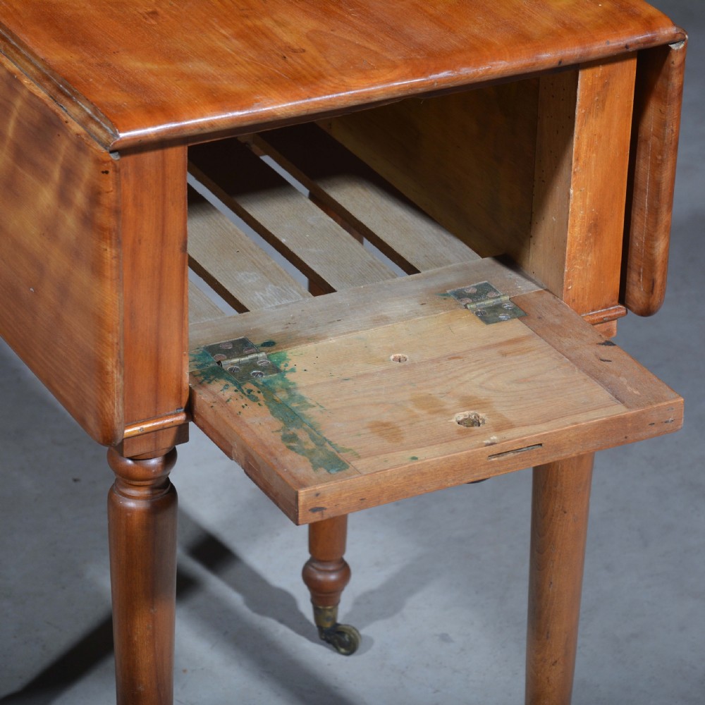 A 19th-century satin birch bedside table featuring a drop-leaf top, two small drawers with dark circular knobs, and turned legs terminating in brass castors. View VII