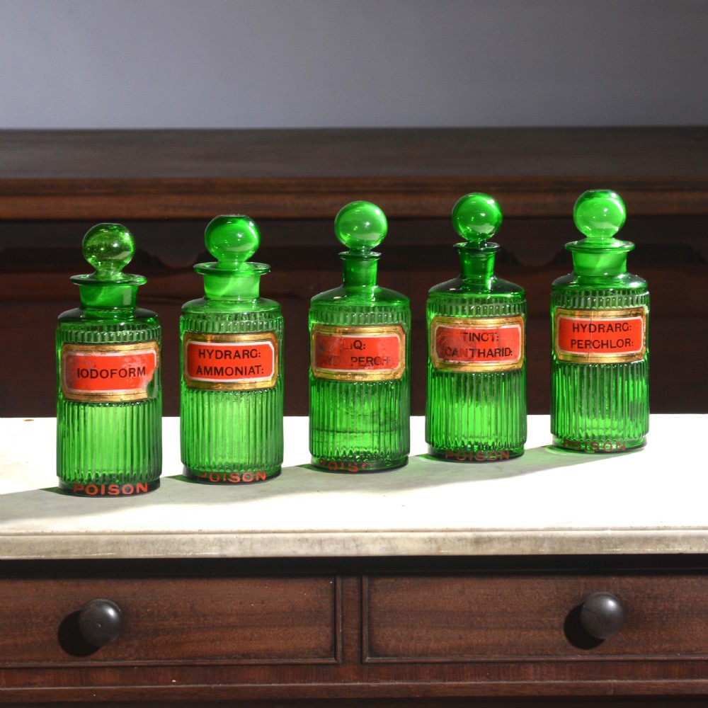 A row of five ribbed emerald green glass apothecary bottles with ground glass stoppers and red gilt-edged labels.