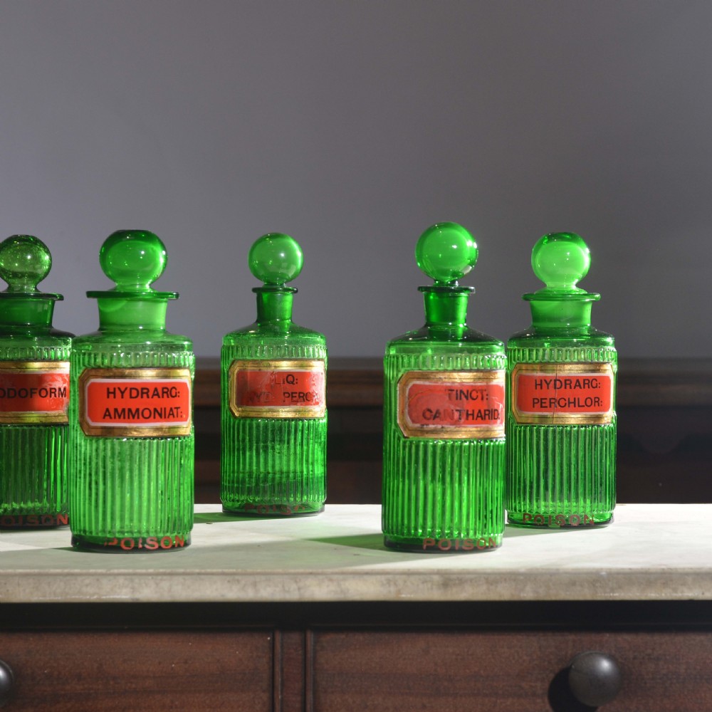 A row of five ribbed emerald green glass apothecary bottles with ground glass stoppers and red gilt-edged labels. View II