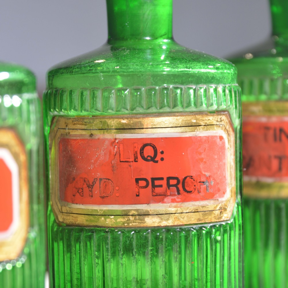 A row of five ribbed emerald green glass apothecary bottles with ground glass stoppers and red gilt-edged labels. View III