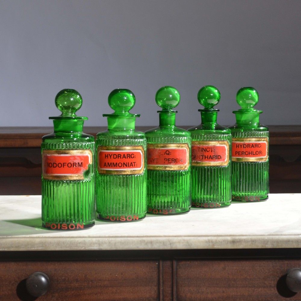 A row of five ribbed emerald green glass apothecary bottles with ground glass stoppers and red gilt-edged labels. View IV