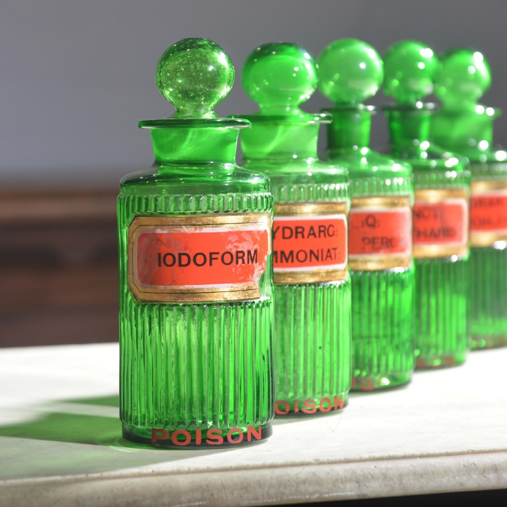 A row of five ribbed emerald green glass apothecary bottles with ground glass stoppers and red gilt-edged labels. View V