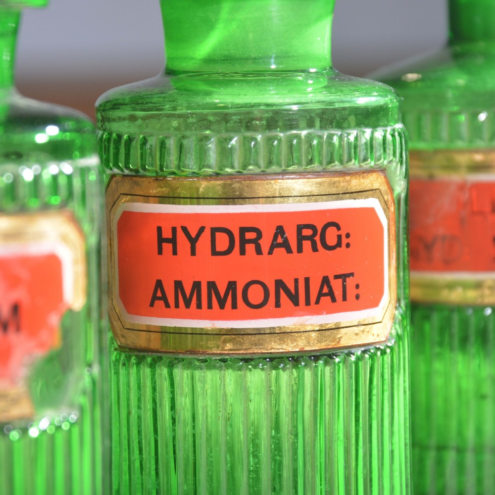 A row of five ribbed emerald green glass apothecary bottles with ground glass stoppers and red gilt-edged labels. View VI