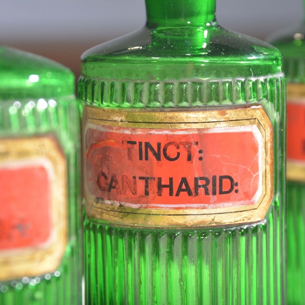 A row of five ribbed emerald green glass apothecary bottles with ground glass stoppers and red gilt-edged labels. View VII