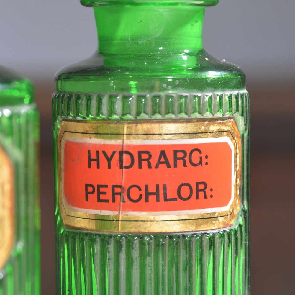 A row of five ribbed emerald green glass apothecary bottles with ground glass stoppers and red gilt-edged labels. View VIII