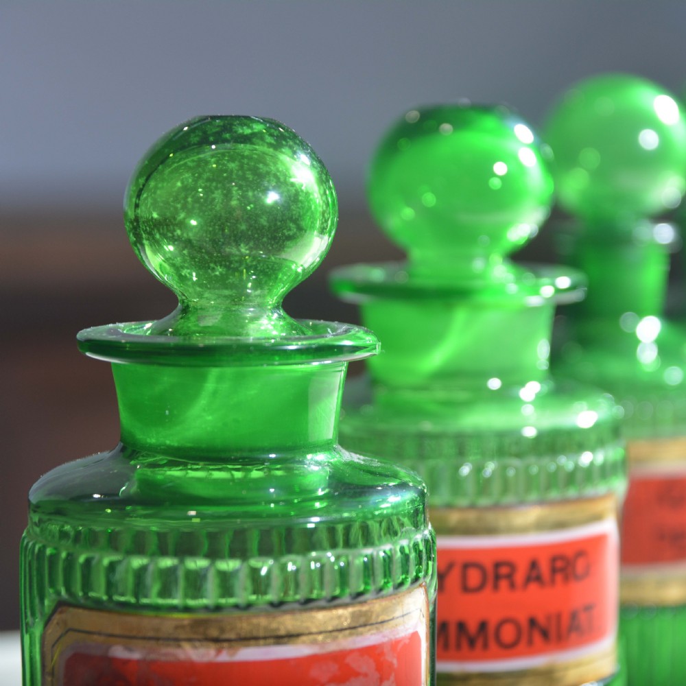A row of five ribbed emerald green glass apothecary bottles with ground glass stoppers and red gilt-edged labels. View X