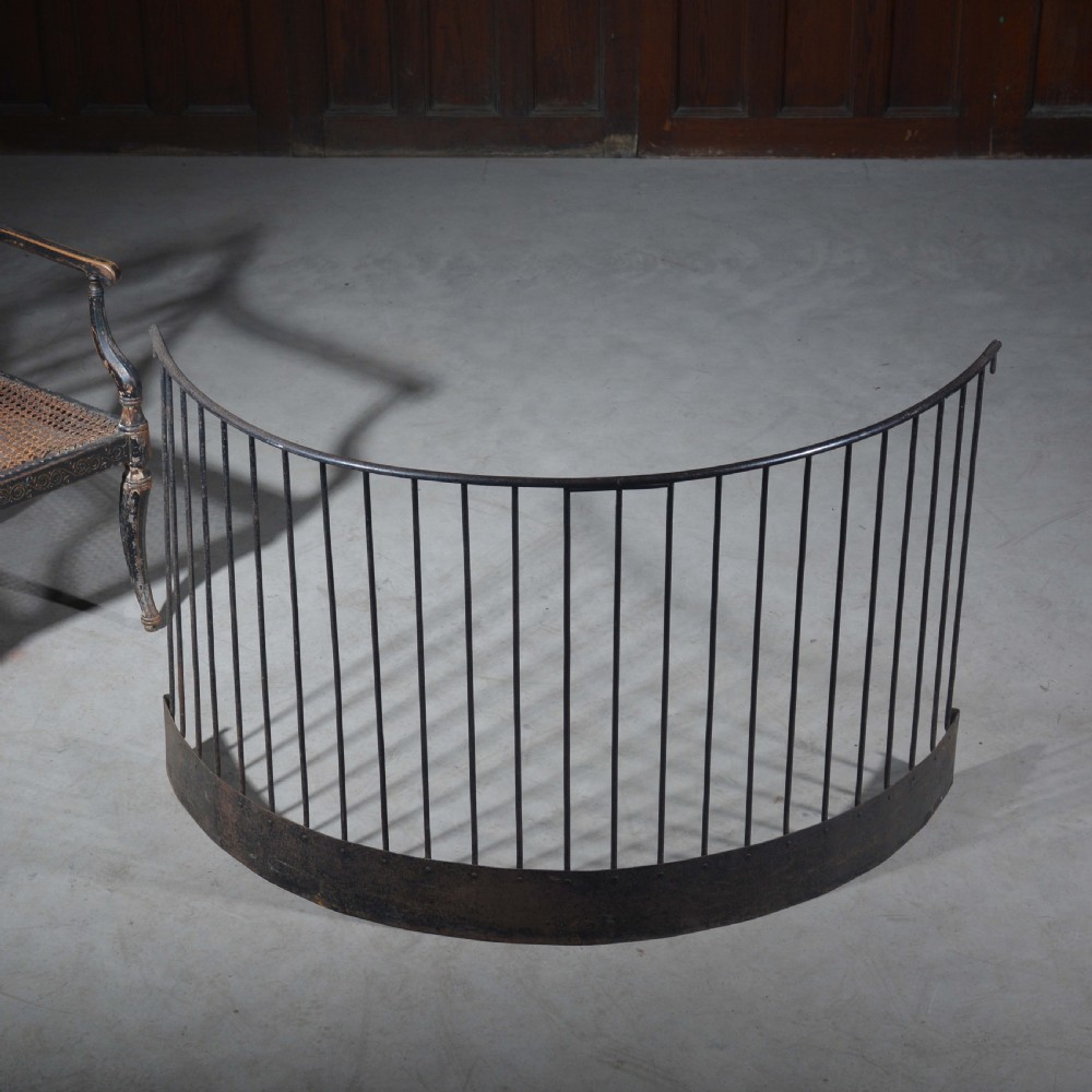 A curved wrought iron nursery fender with closely spaced vertical bars mounted onto a solid metal plinth base.