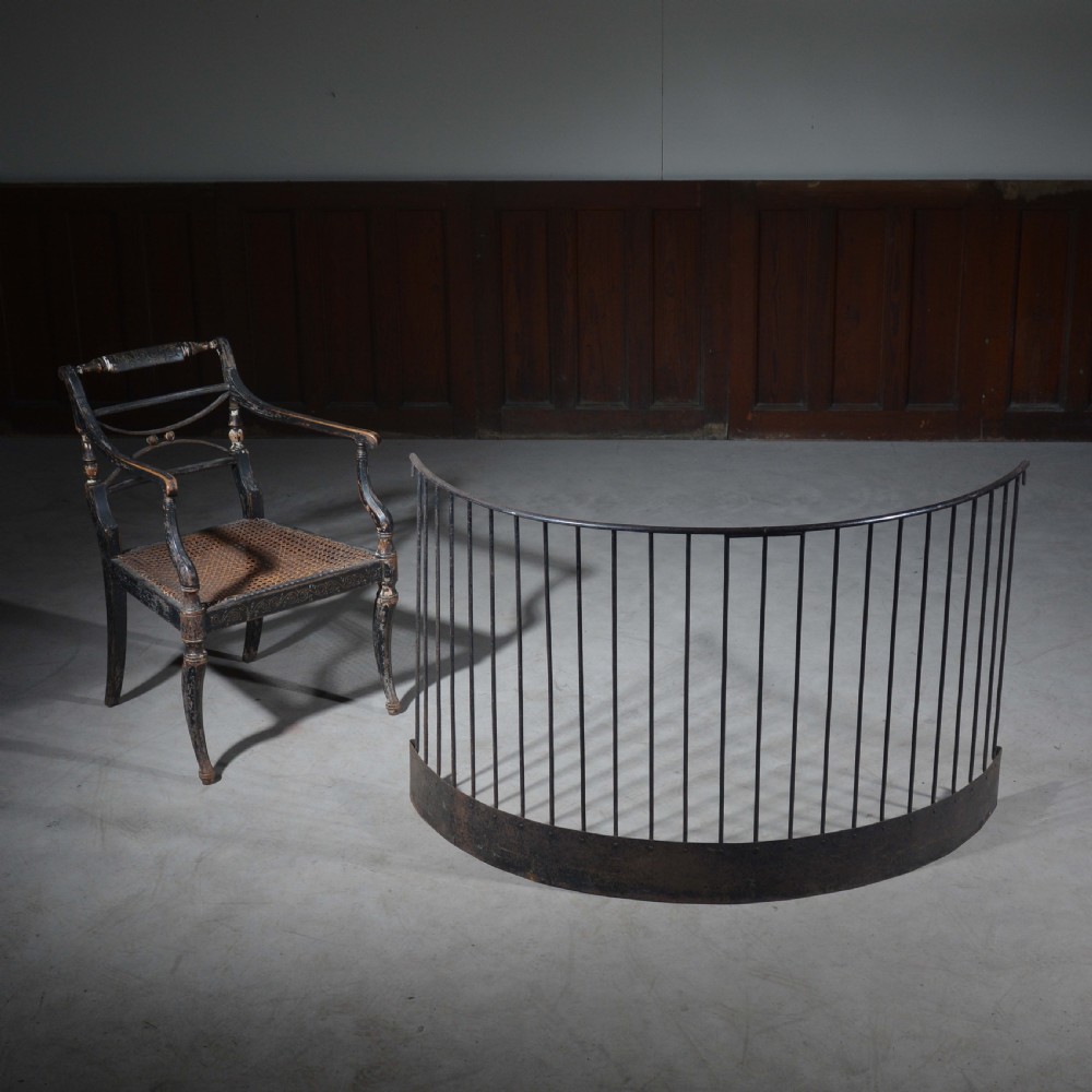 A curved wrought iron nursery fender with closely spaced vertical bars mounted onto a solid metal plinth base. View II