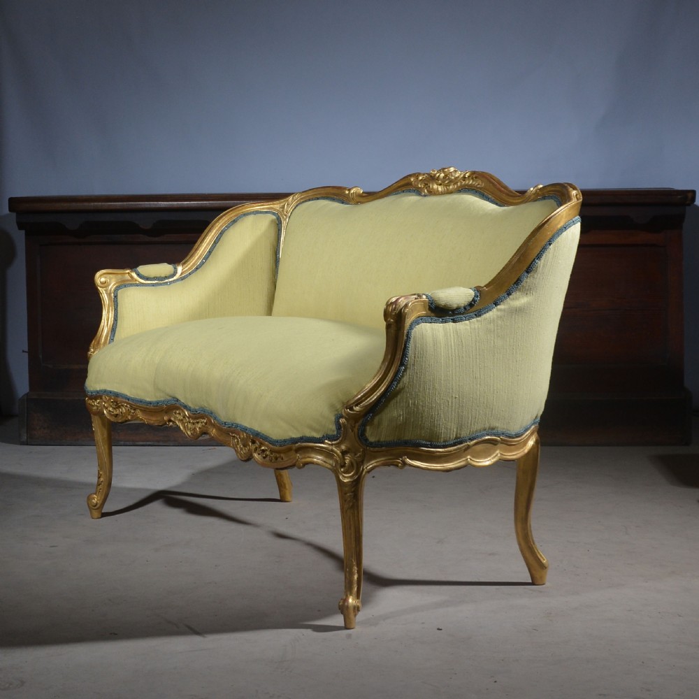 A French Louis XV style giltwood settee featuring a curved carved frame, cabriole legs, and light yellow fabric upholstery with blue trim.