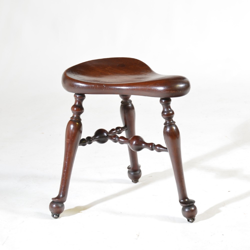 An antique dark walnut three-legged saddle stool featuring a dished seat, turned legs, and decorative spindle stretchers.