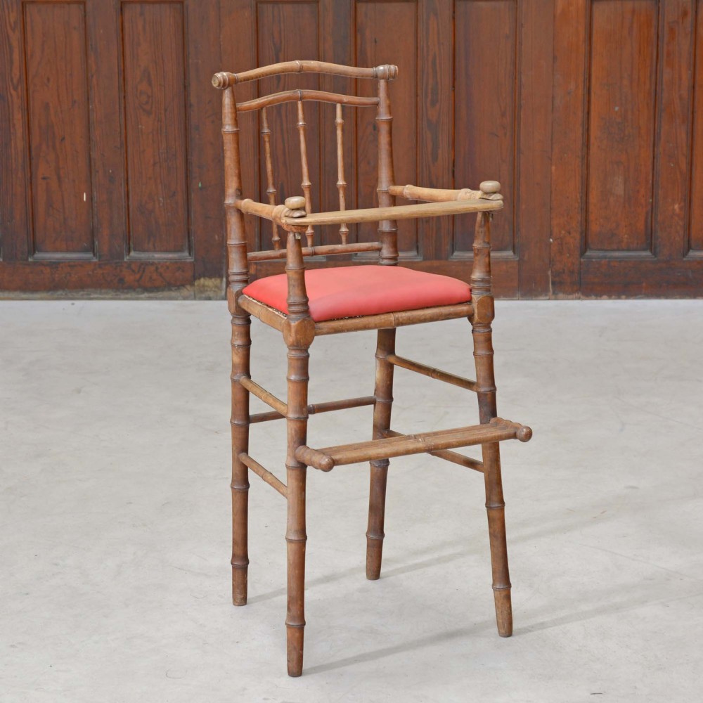 An antique French highchair featuring a turned faux bamboo frame, a red upholstered seat, an integrated safety bar, and a wooden footrest.