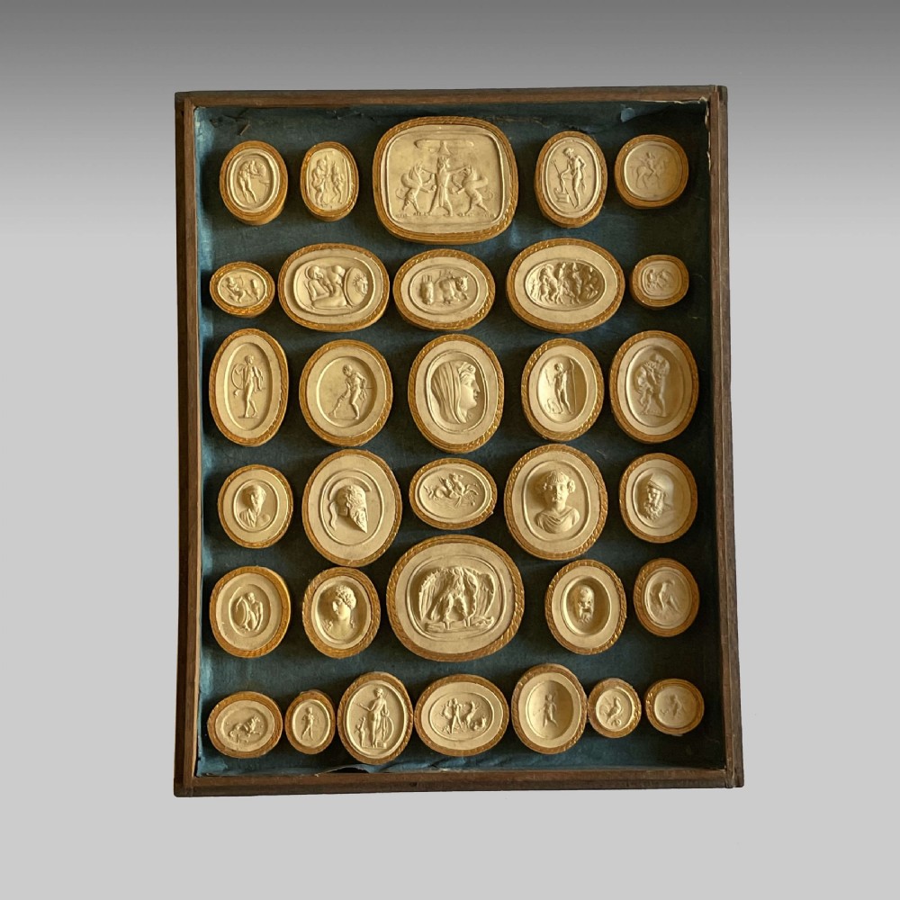 A dark wooden frame containing a teal velvet backing mounted with numerous oval plaster intaglio casts of classical figures framed in gold-toned rims.