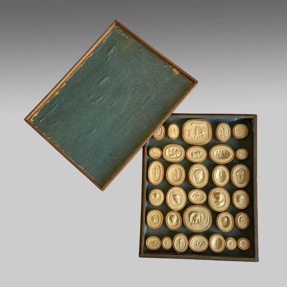 A dark wooden frame containing a teal velvet backing mounted with numerous oval plaster intaglio casts of classical figures framed in gold-toned rims. View II