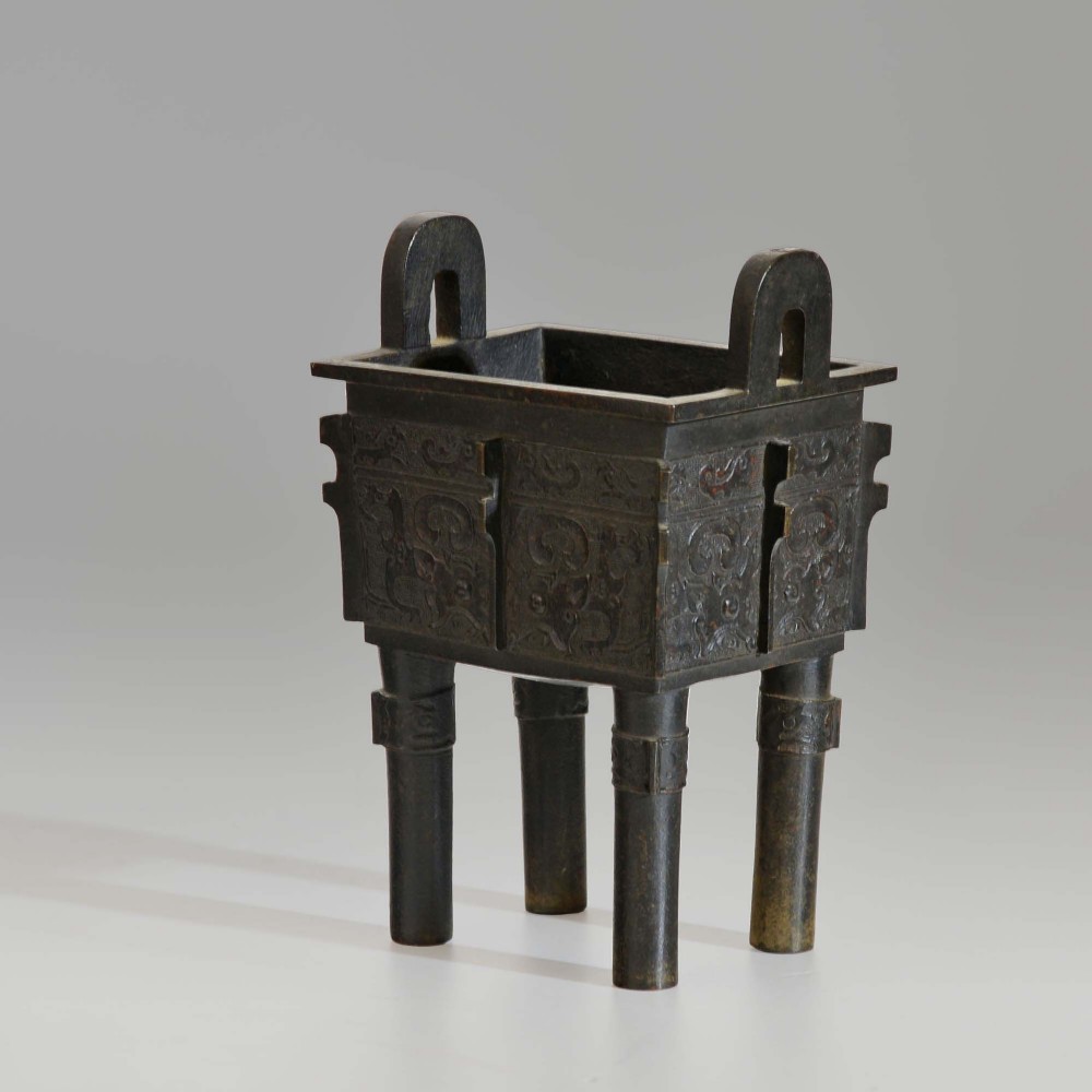 A rectangular dark bronze ritual vessel featuring four straight legs, two upright loop handles, relief carvings, and corner flanges.