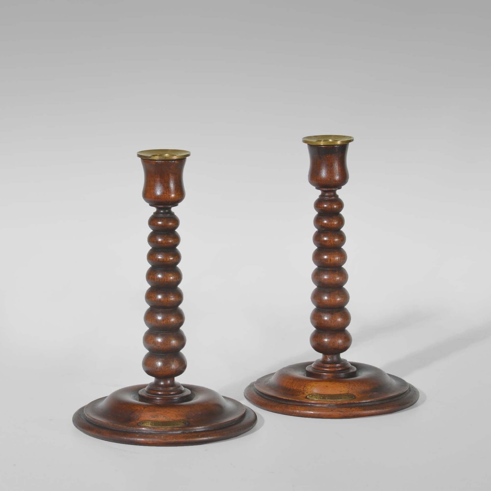 A pair of dark turned teak candlesticks featuring repeating bobbin stems, circular bases with small brass plaques, and brass sconce inserts at the top.